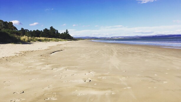 Une vaste plage de sable fin parsemée d’empreintes, un ciel bleu azur et des montagnes lointaines.