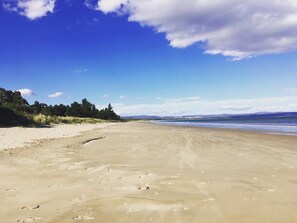 Una amplia playa arenosa con huellas, cielo azul despejado y montañas a lo lejos.