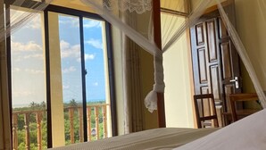 A hotel room with a large bed, a balcony with a view of greenery and the sea, and a wooden chair. - Authentic Mikoko Beach & Cottages (Bagamoyo)