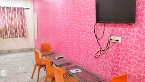 A room with a pink patterned wall, a TV mounted on the wall, and a table with chairs. - Hotel Ram Palace (New Delhi)