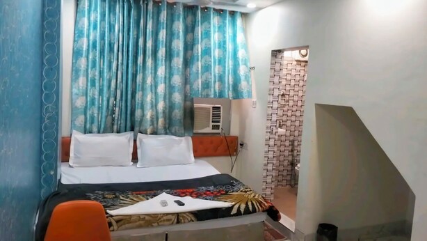 A bedroom with a bed, pillows, a bedside table, an air conditioning unit, and a patterned wall. - Hotel Ram Palace (New Delhi)