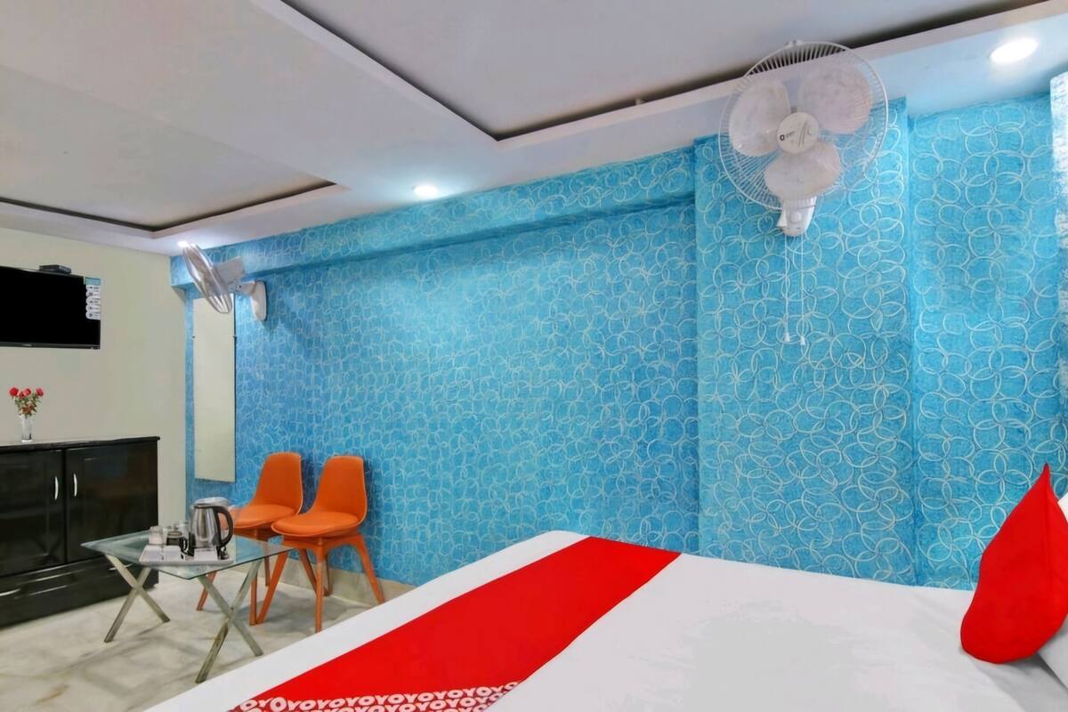 Habitación de hotel con pared azul estampada, cama con colcha roja y blanca, dos sillas naranjas, una mesita con tetera y un ventilador de techo.