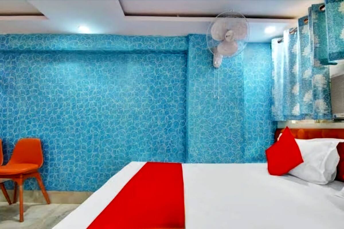 Habitación de hotel con cama, colcha roja y blanca, pared con estampado azul, ventilador y una silla naranja.