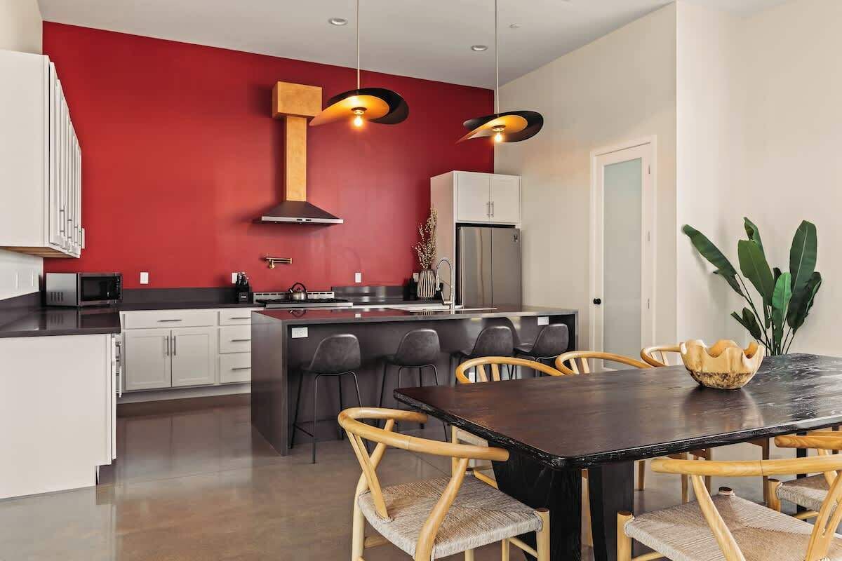 Una cocina moderna con una pared roja de acento, una isla central con taburetes y una zona de comedor con mesa oscura y sillas de mimbre.