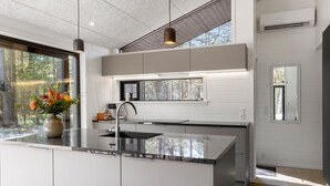 A modern kitchen with a large island, white cabinetry, a built-in oven, and a window overlooking a forest.