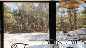 A dining area with a table and chairs, overlooking a forested area with a dirt path.