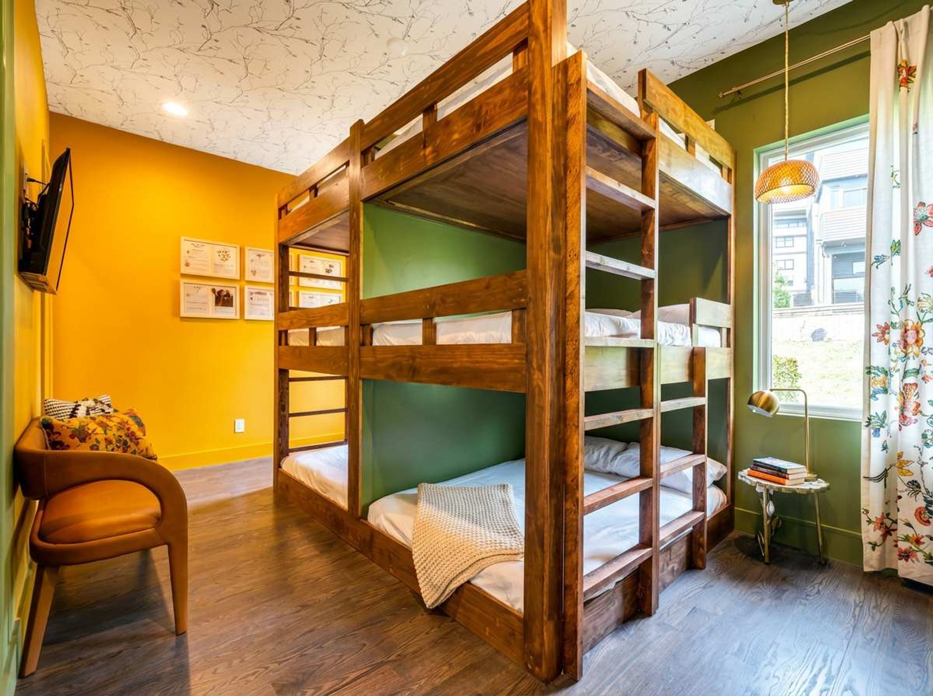 A bunk bed room with a wooden frame, a chair, a small table with books, and a window with curtains.
