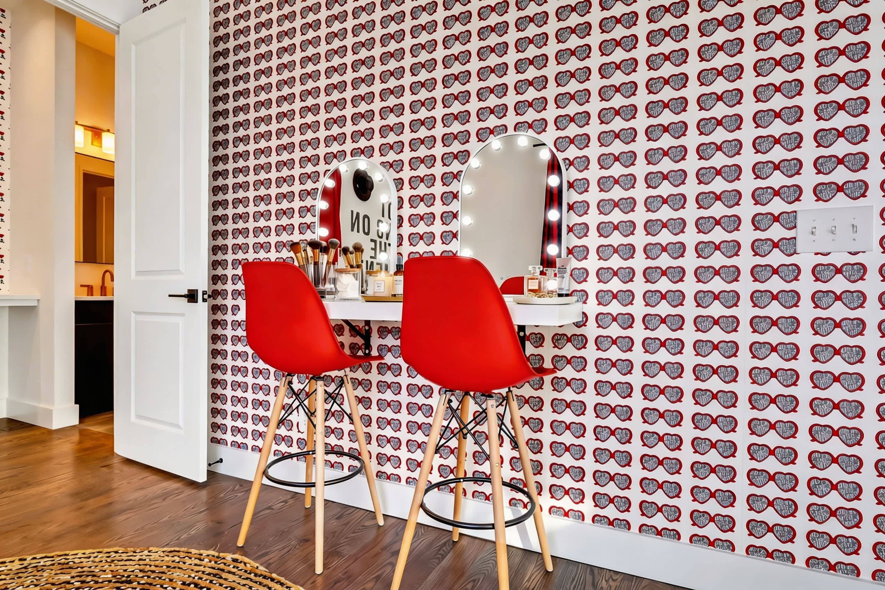 A room with a vanity table, two red chairs, a mirror, and a patterned wallpaper.