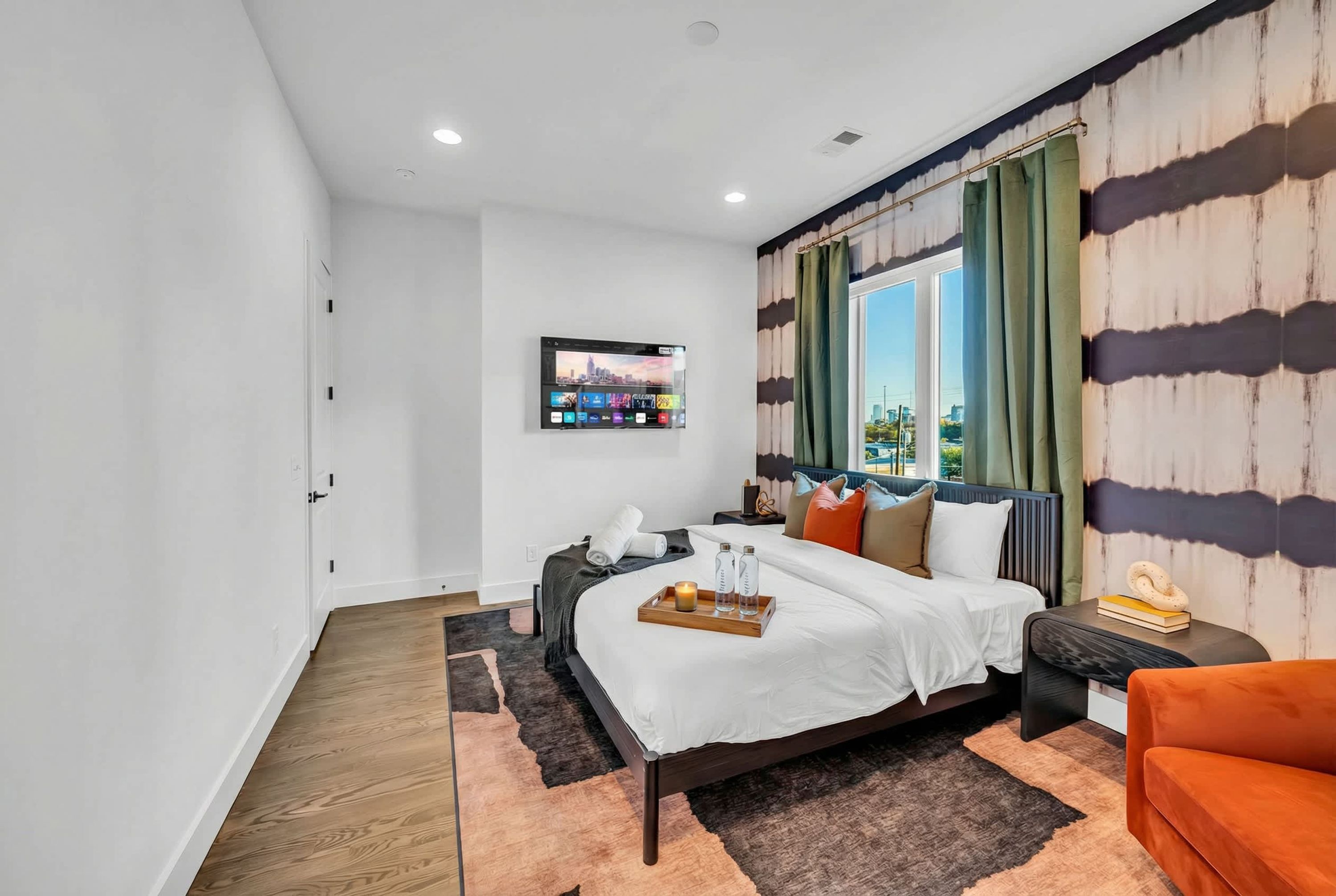 A modern bedroom with a bed, a TV mounted on the wall, a window with curtains, and a view of the cityscape.