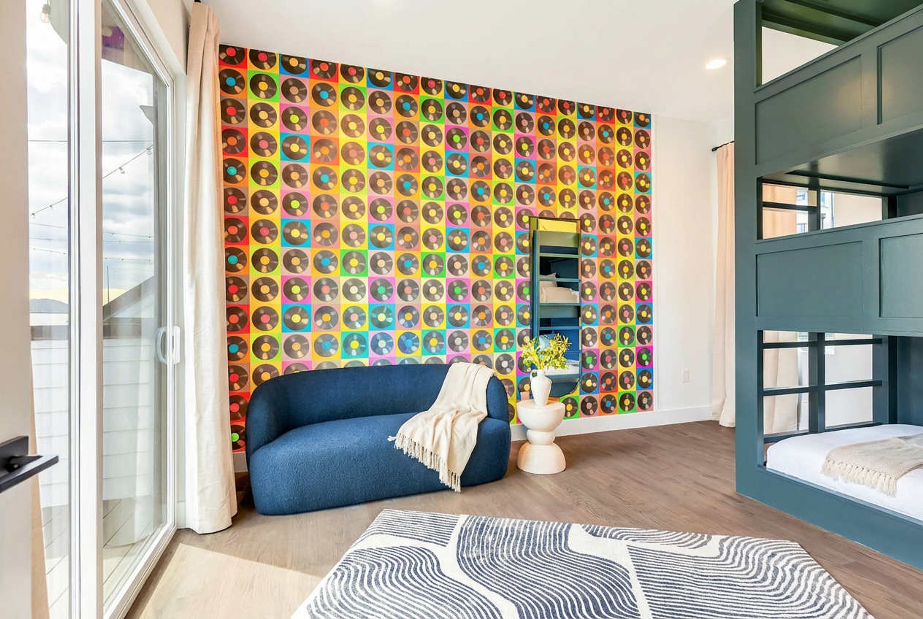 A room with a colorful vinyl record wall art, a blue sofa, a white bedside table, and a bunk bed.