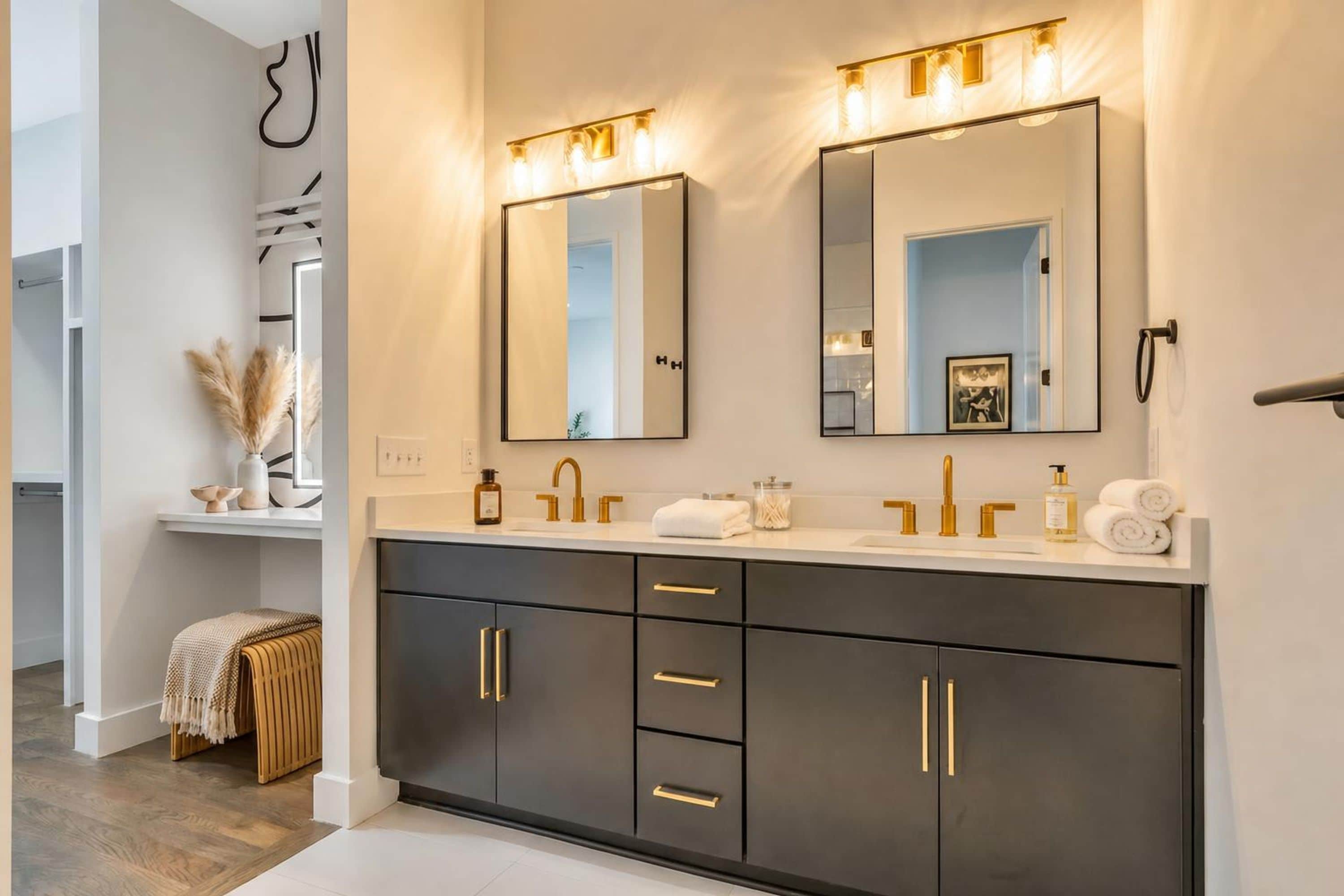 Un baño moderno con un mueble de tocador oscuro, pomos dorados y un espejo grande con iluminación.