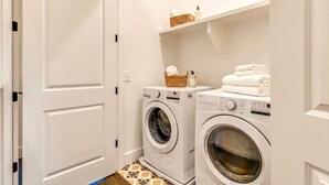 A laundry room with two front-loading washing machines, a shelf with towels and a basket, and a door with a handle. - New Hot Tub Rooftop Decks City Views Poker (Nashville)