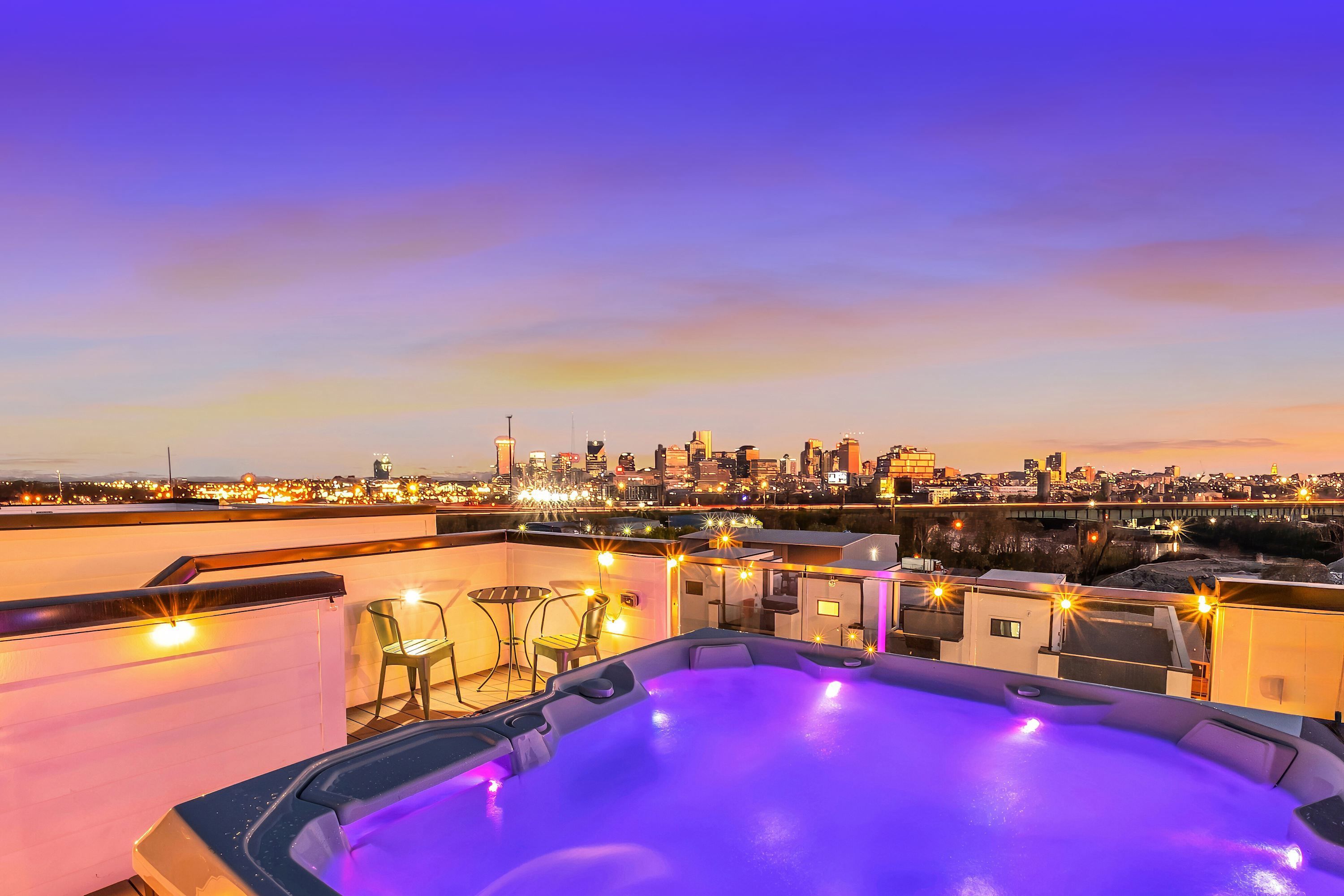 Una terraza en la azotea con jacuzzi, mobiliario exterior y vista al horizonte urbano al anochecer.