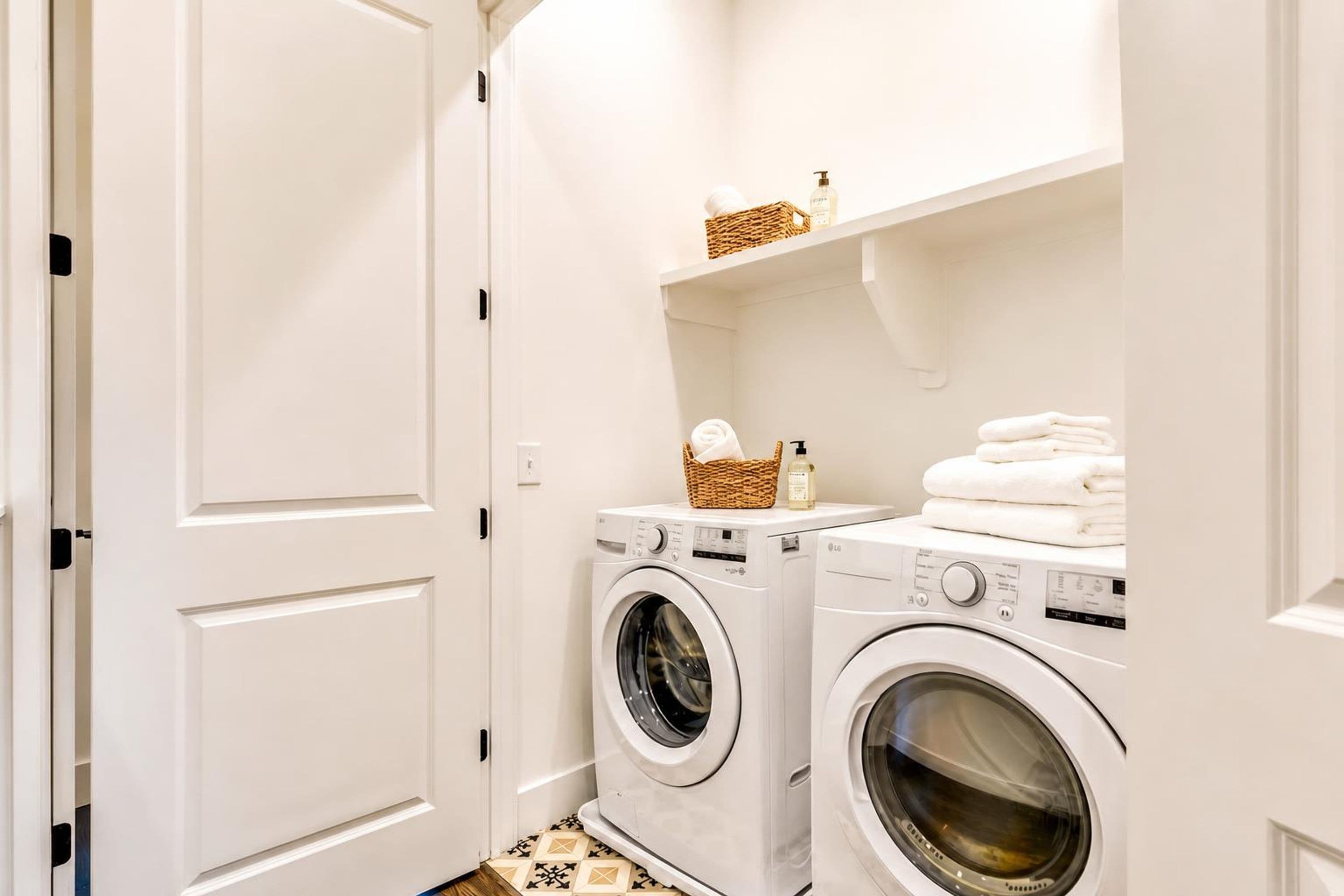 A compact laundry room with two front-loading washing machines, a shelf with folded towels, and a basket with a bottle.