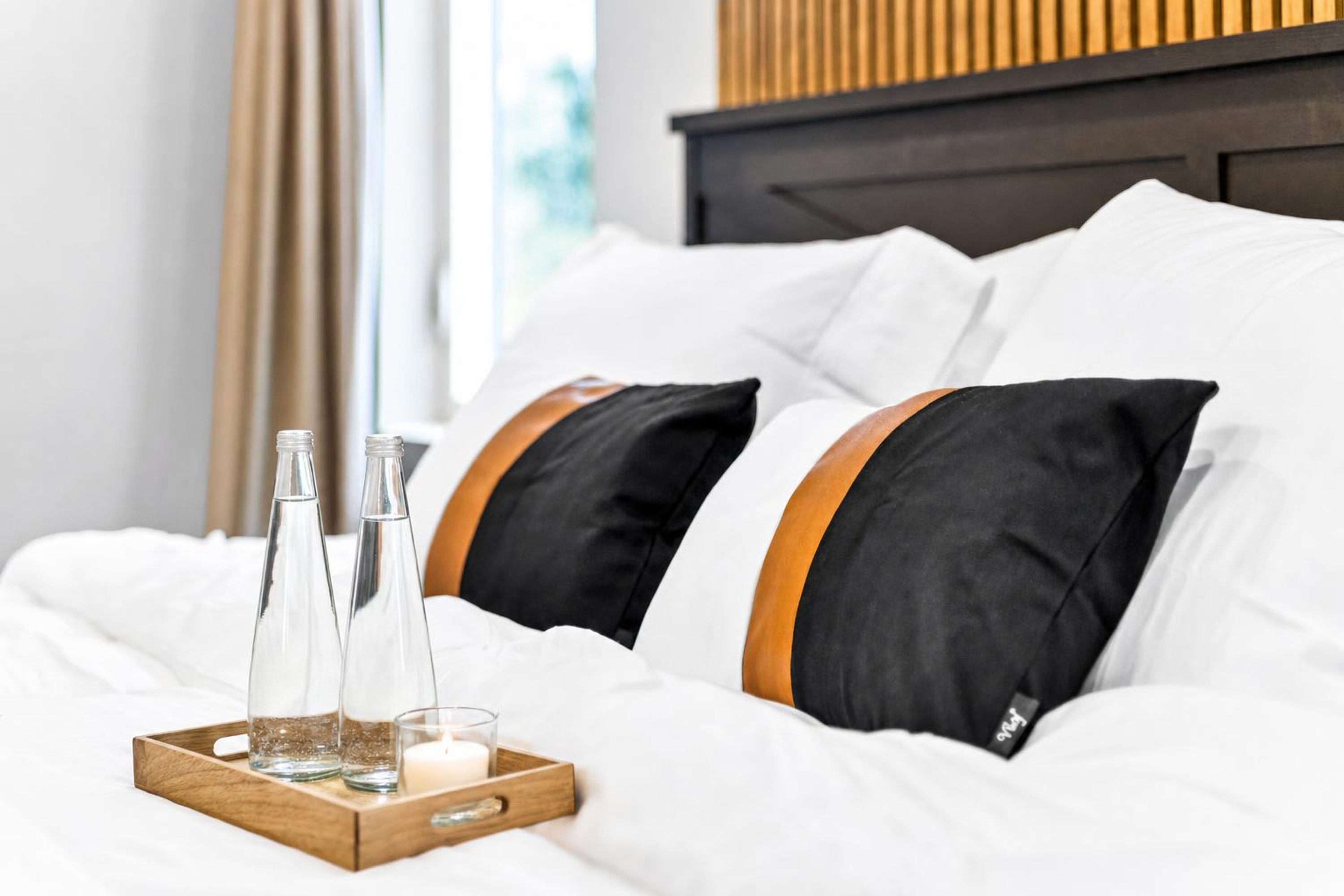 Una cama bien hecha con ropa blanca, dos almohadas negras con rayas anaranjadas y una bandeja de madera con dos botellas transparentes y una vela.