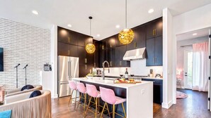 Une cuisine moderne avec des meubles de couleur sombre, un îlot central blanc, des tabourets de bar roses et des luminaires suspendus.