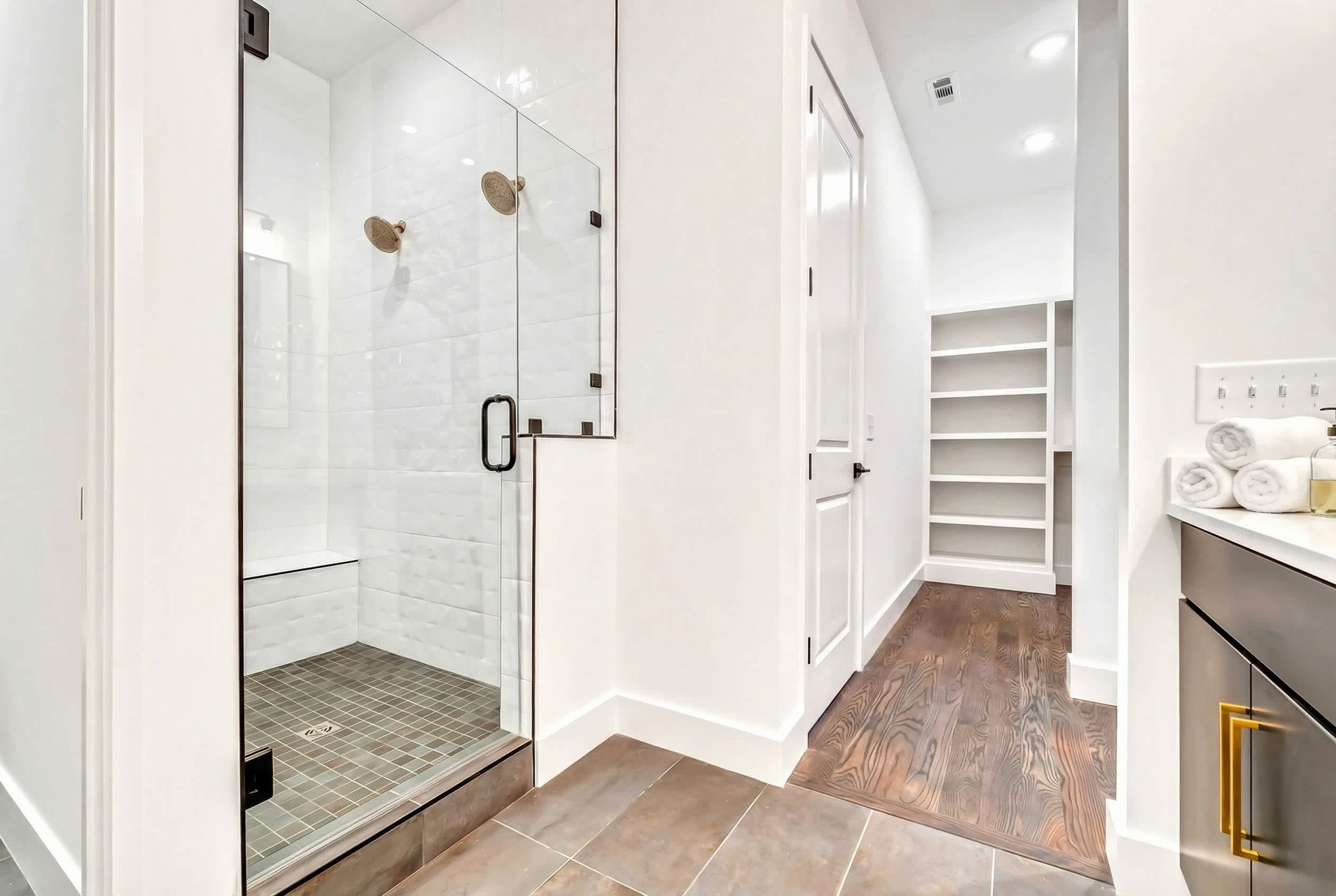 A modern bathroom with a glass shower enclosure, a walk-in closet with shelves, and a wooden floor.