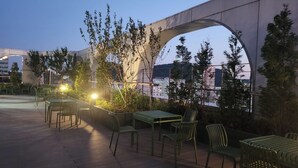 Een terras met tafels en stoelen, omringd door groen en met uitzicht op een stadsbeeld.