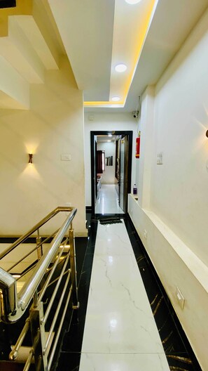 Un pasillo moderno de hotel con suelo de mármol, barandales metálicos, iluminación empotrada en el techo y un extintor en la pared.