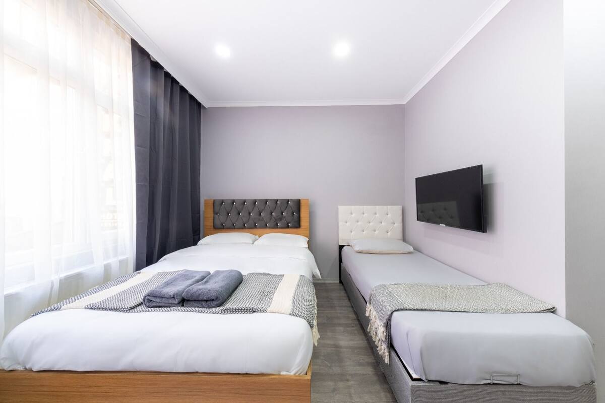 Ein Hotelzimmer mit einem großen Bett, einem kleineren Bett, einem Fernseher und einem Fenster mit Vorhängen.