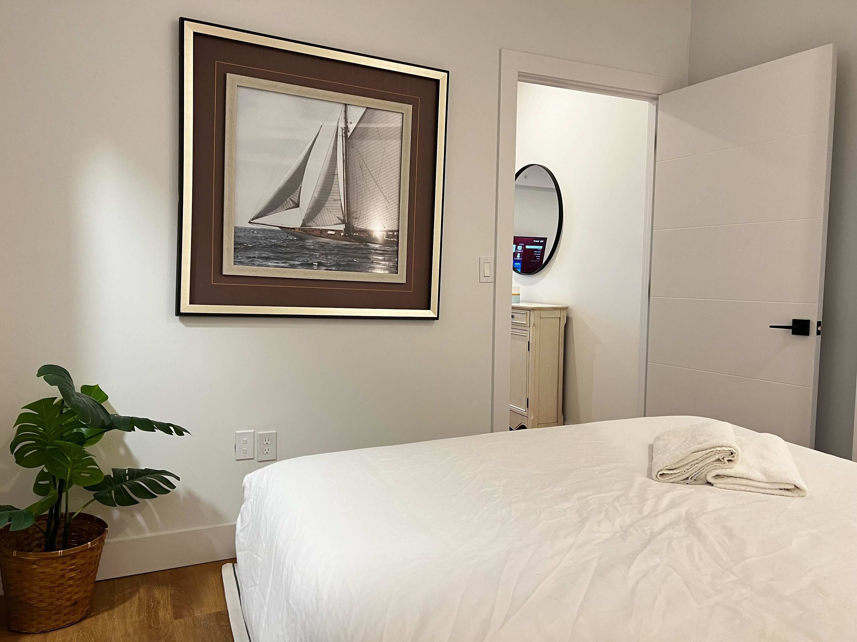 A hotel room with a bed, a framed picture of a sailboat, a potted plant, a mirror, and a door.