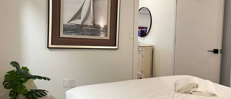 A hotel room with a bed, a framed picture of a sailboat, a potted plant, a mirror, and a door.