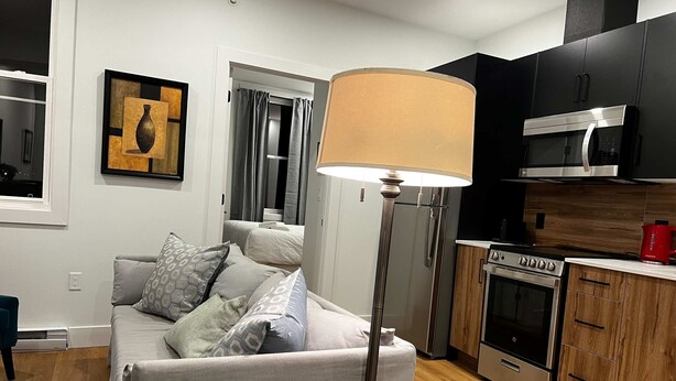 Una sala de estar moderna con sof á, lámpara, cocina equipada y una puerta que da a otra habitación.