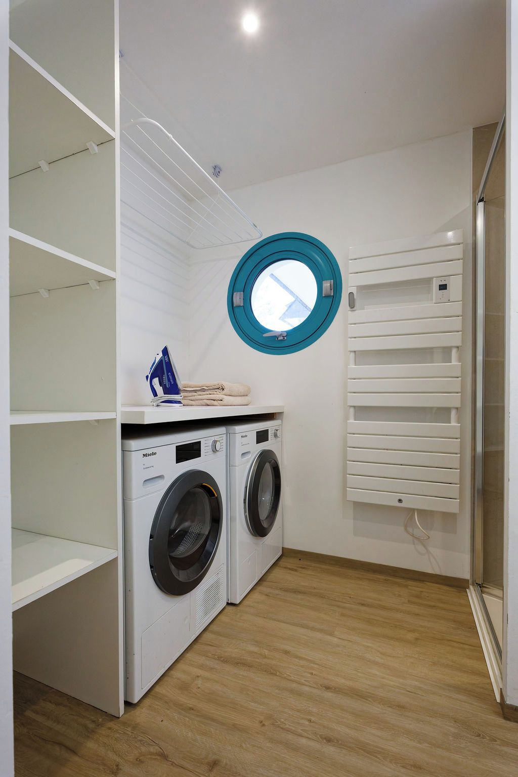 Une buanderie avec deux machines à laver blanches, une fenêtre bleue de forme circulaire et un étendoir à linge blanc.