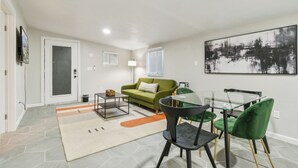 A modern living room with a glass dining table, green and black chairs, a green sofa, and a large abstract painting on the wall.