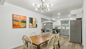A modern kitchen with white cabinets, a stainless steel refrigerator, and a dining area with a wooden table and white chairs. - Pool & Games | Peaceful Neighborhood (Phoenix)