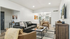 A modern living room with a black leather sofa, a brown leather armchair, and a coffee table. There is a dining area with a white table and chairs in the background. - Pool & Games | Peaceful Neighborhood (Phoenix)