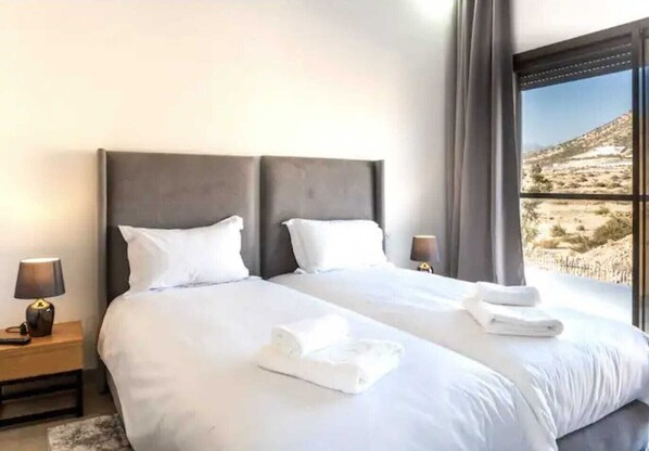 Una cama doble con sábanas blancas y dos almohadas, una mesita de noche con lámpara, y una ventana con vistas a un paisaje desértico.