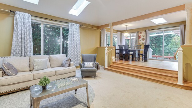 Un vaste salon comprenant un canapé, un fauteuil et une table basse, de grandes fenêtres et un escalier menant à une salle à manger.