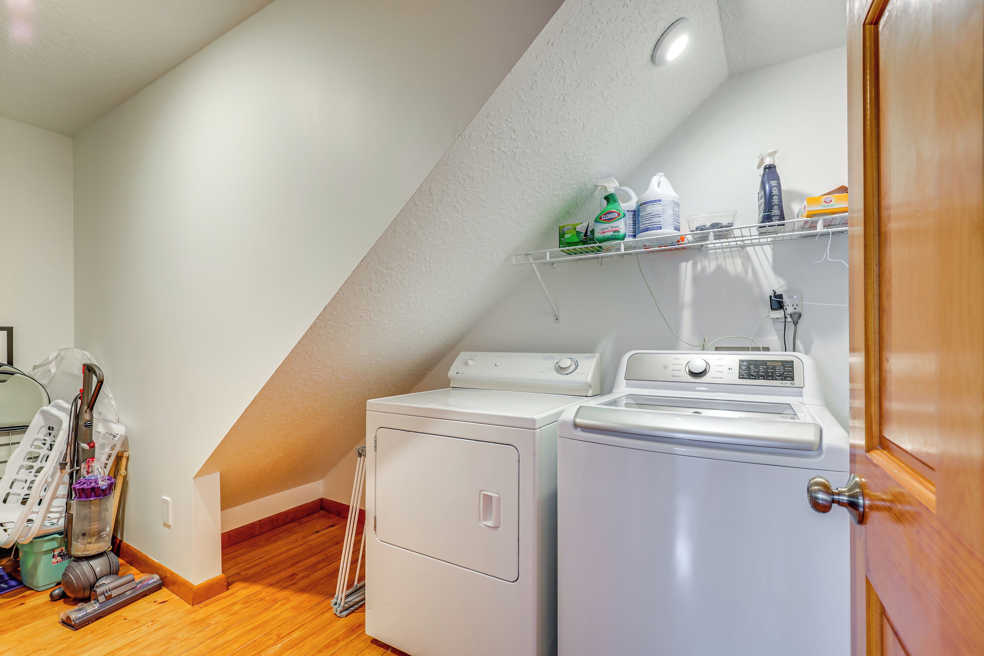 A laundry room with two white washing machines, a shelf with cleaning supplies, and a vacuum cleaner.