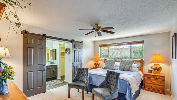 A bedroom with a bed, two chairs, a nightstand, a lamp, a ceiling fan, and a sliding door leading to another room. - Cyclists' Haven: Southwestern-style Home in Tucson (Tucson)