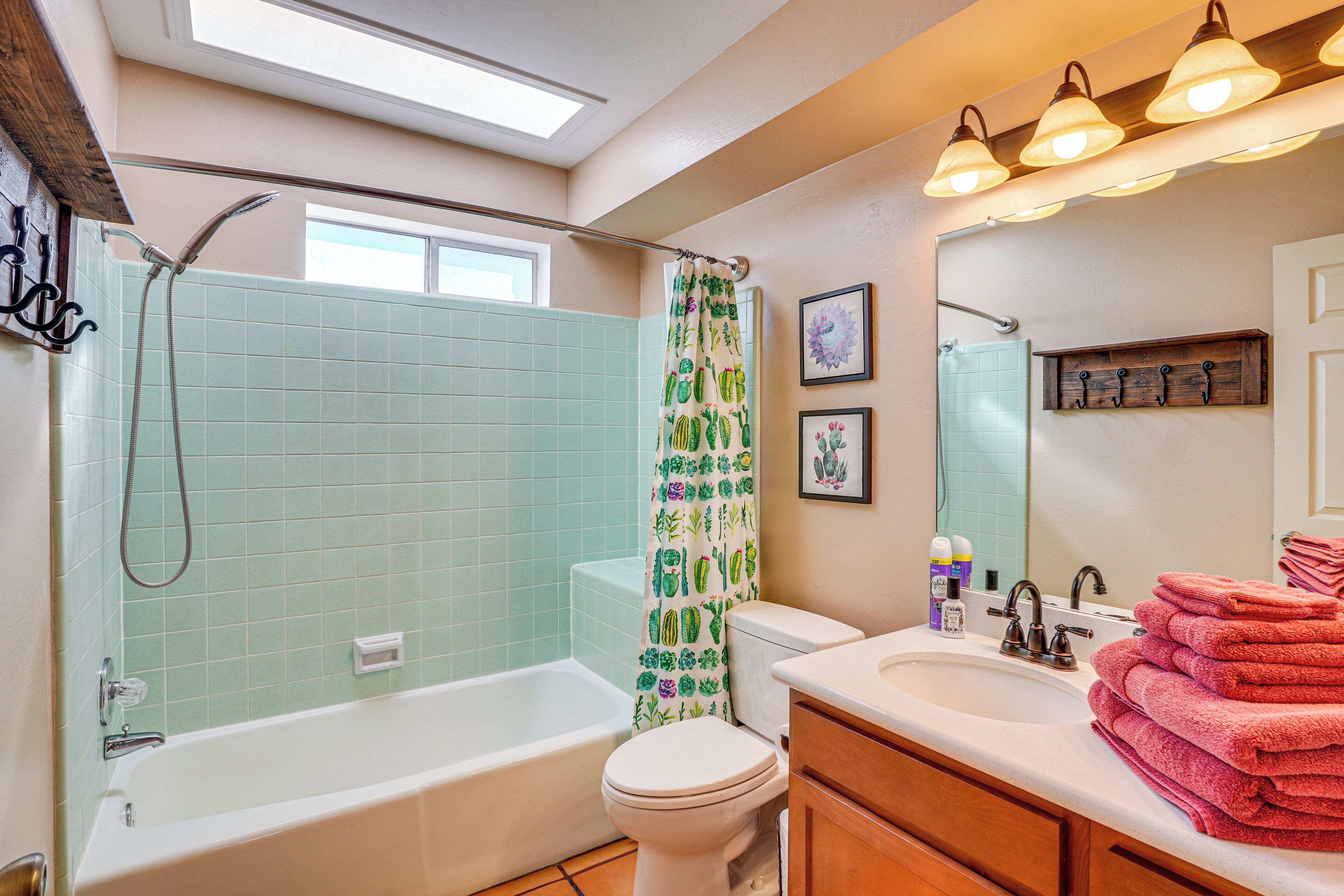 Une salle de bain dotée d’une lucarne, d’une douche avec un rideau orné de motifs de cactus, de toilettes, d’un lavabo avec un miroir et d’une pile de serviettes.