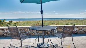A round metal table with two chairs and a large blue umbrella, set against a stone wall with a view of the sea. - 'beachfront 15' Condo on Old Orchard Beach (Old Orchard Beach)