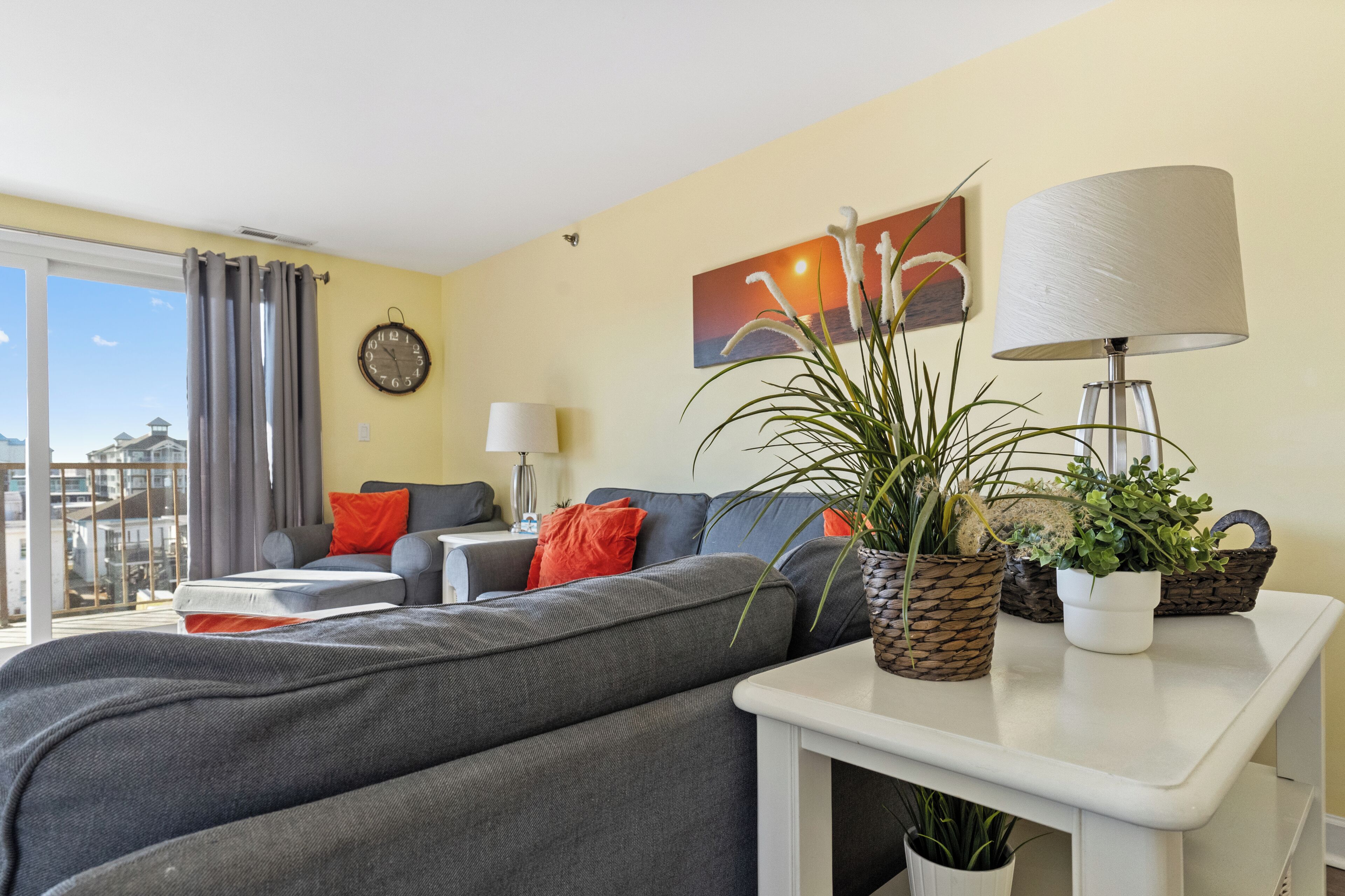 Un salon moderne avec un canapé gris, des coussins orange, une table d’appoint blanche agrémentée de plantes, une lampe et un tableau représentant un coucher de soleil.