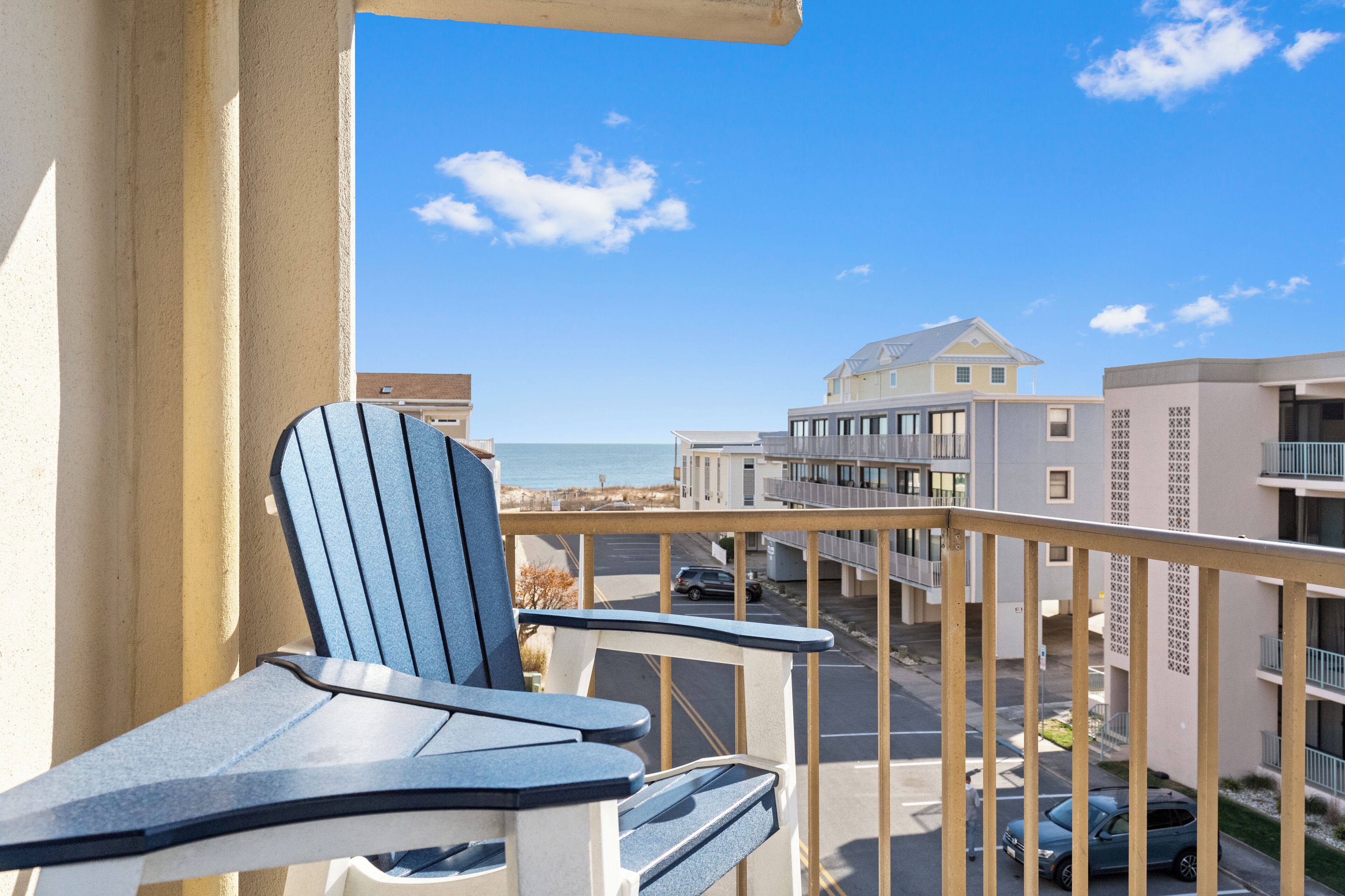 A balcony with a view of the beach and ocean, a chair, and a building with balconies.