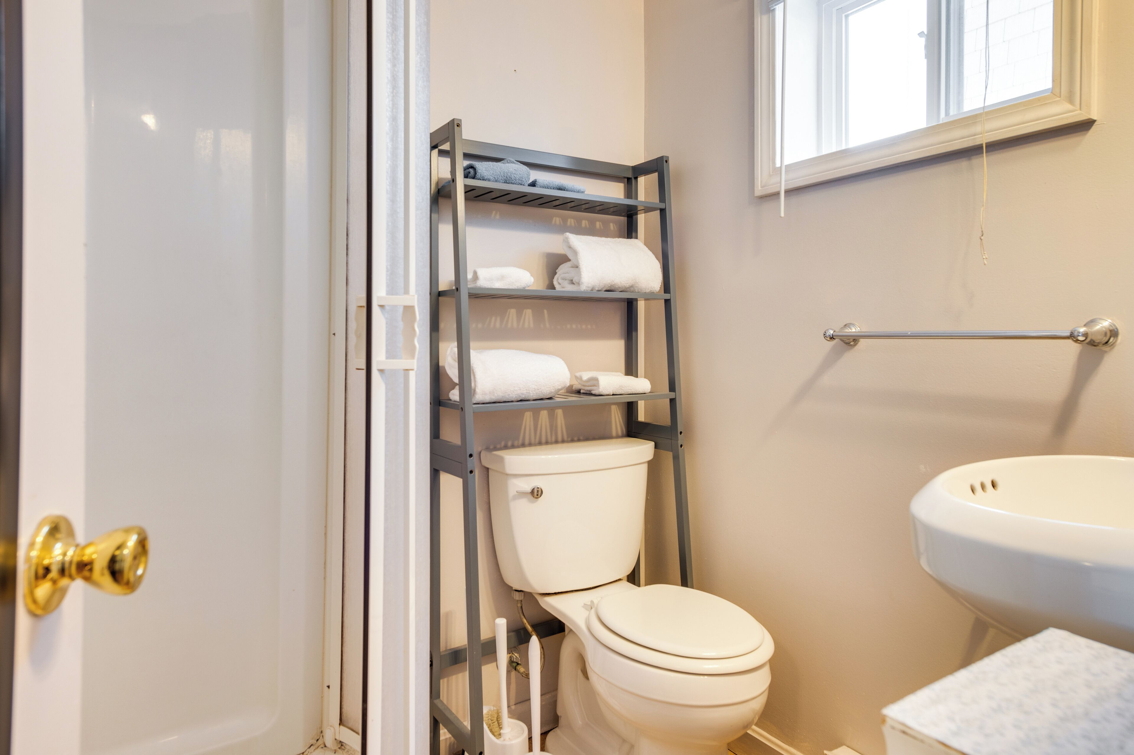 A bathroom with a white toilet, a metal shelf with towels, a window, and a white sink.