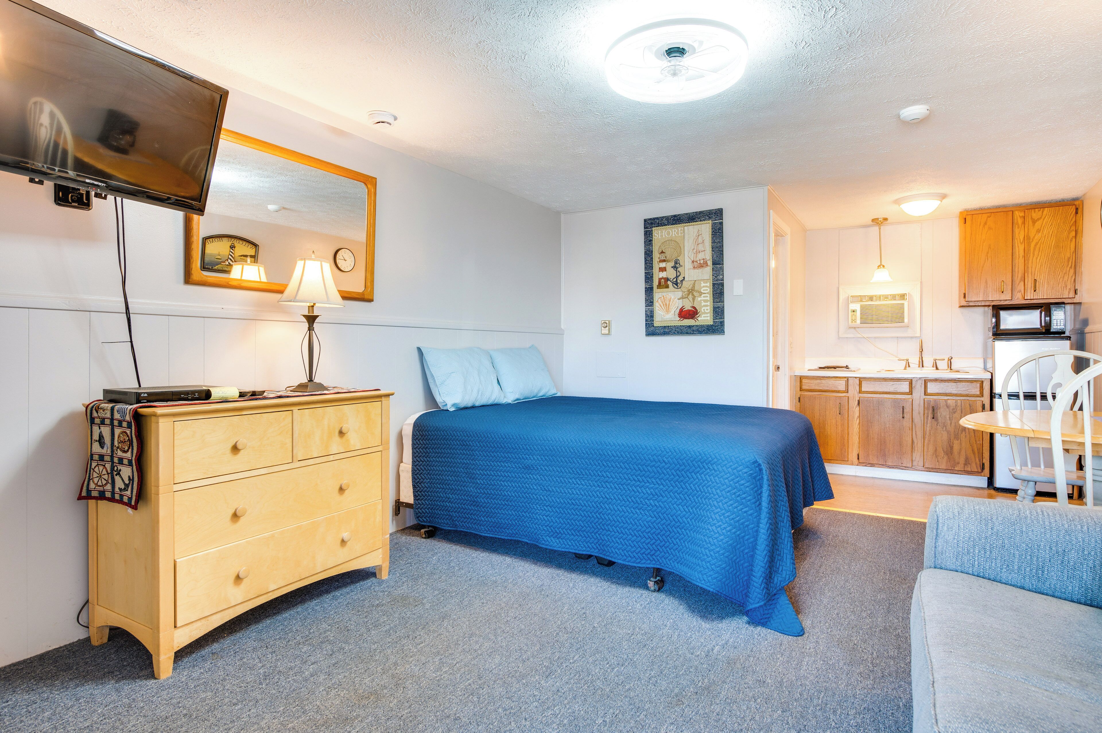 A bedroom with a bed, a dresser, a TV, a lamp, and a kitchen area with cabinets and a microwave.