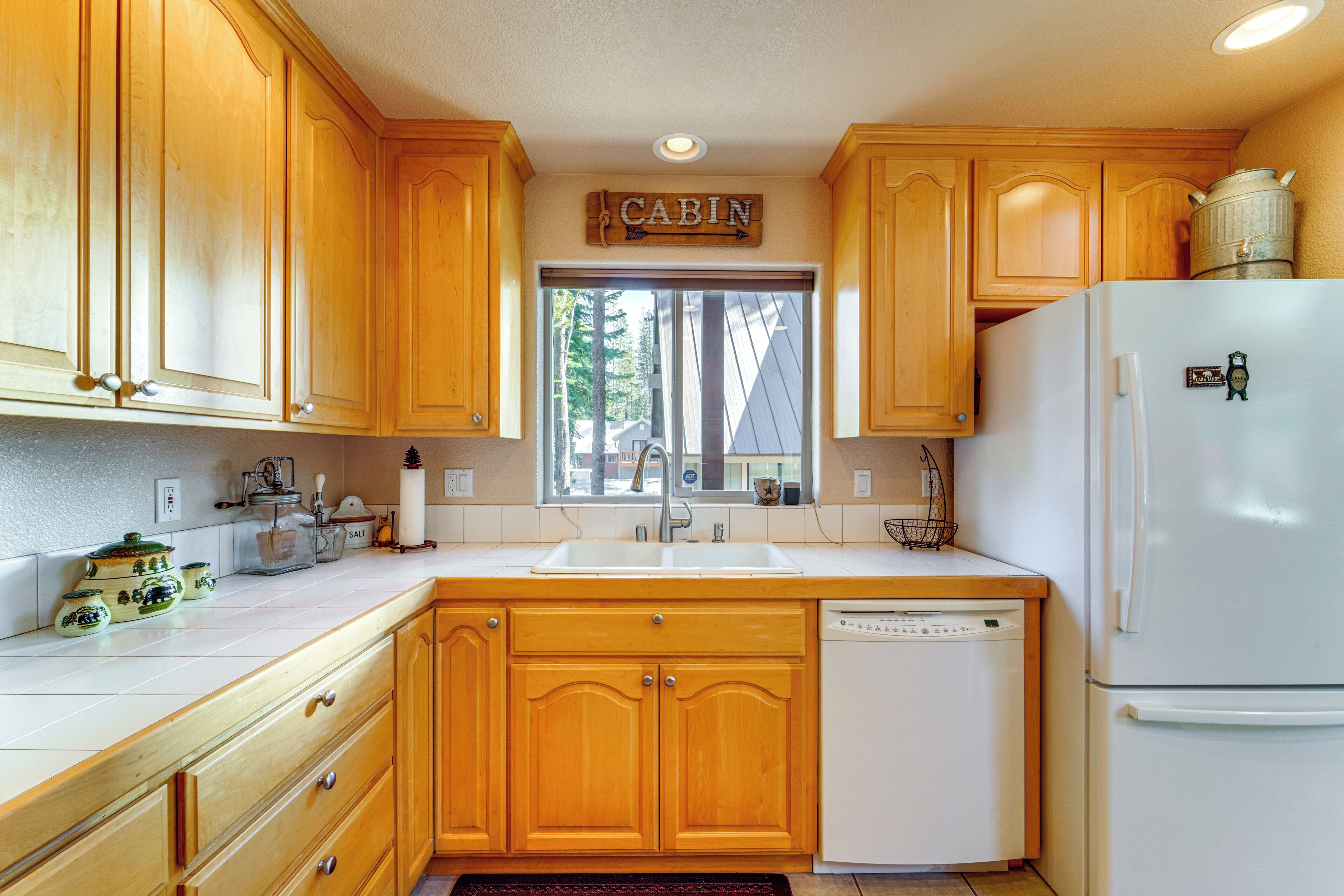 A kitchen with wooden cabinets, a white refrigerator, a dishwasher, and a window with a view of trees.