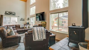 A cozy living room with a brown leather sofa, a black wood-burning stove, and large windows offering a view of a wooden deck and a house. - Neighborhood Beach Access! Soda Springs Getaway (Truckee)
