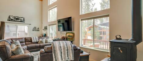 A cozy living room with a brown leather sofa, a black wood-burning stove, and large windows offering a view of a wooden deck and a house.