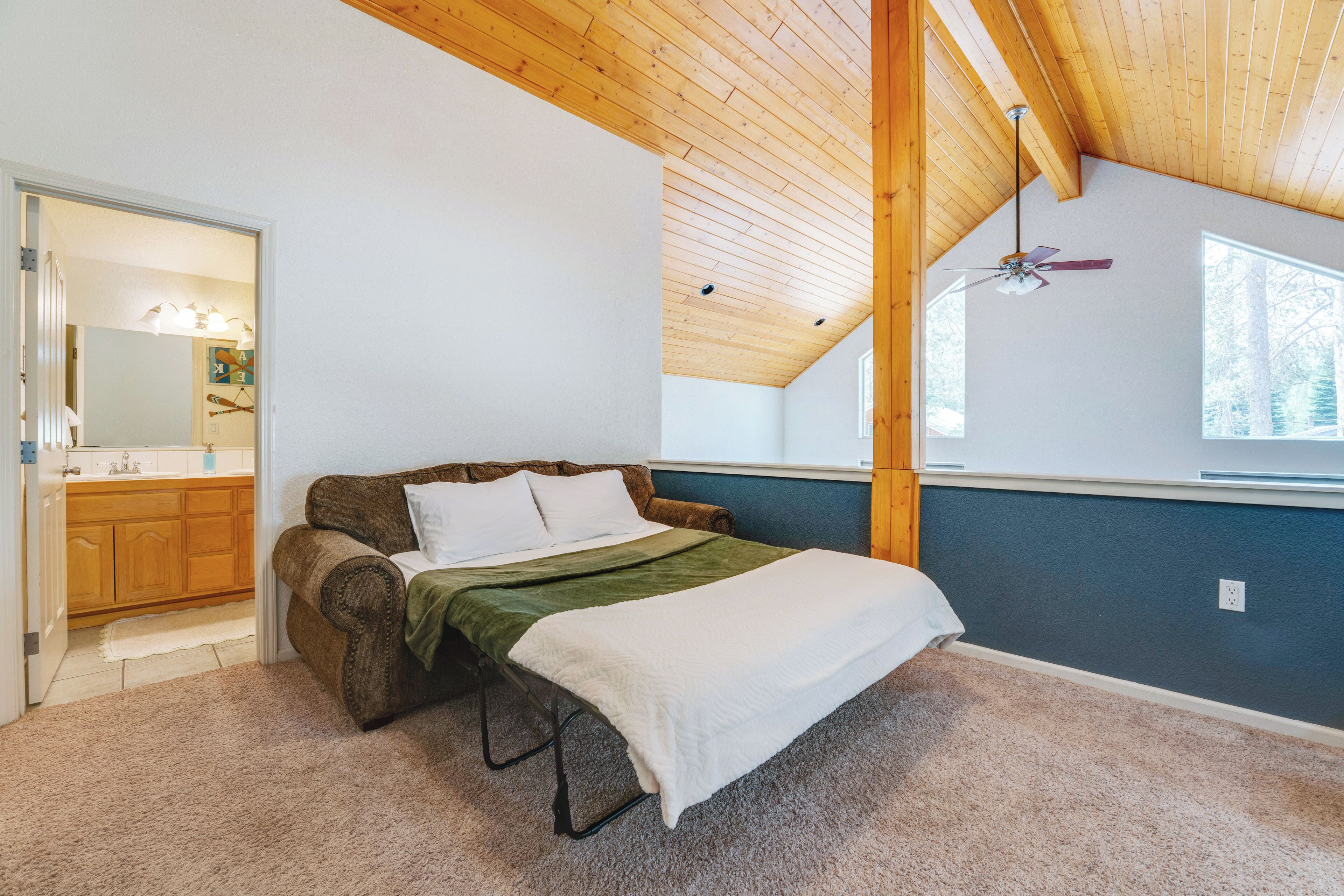 Une chambre avec un plafond de style industriel, un lit recouvert d’une couverture verte, un ventilateur de plafond et une vue sur l’extérieur par une fenêtre.