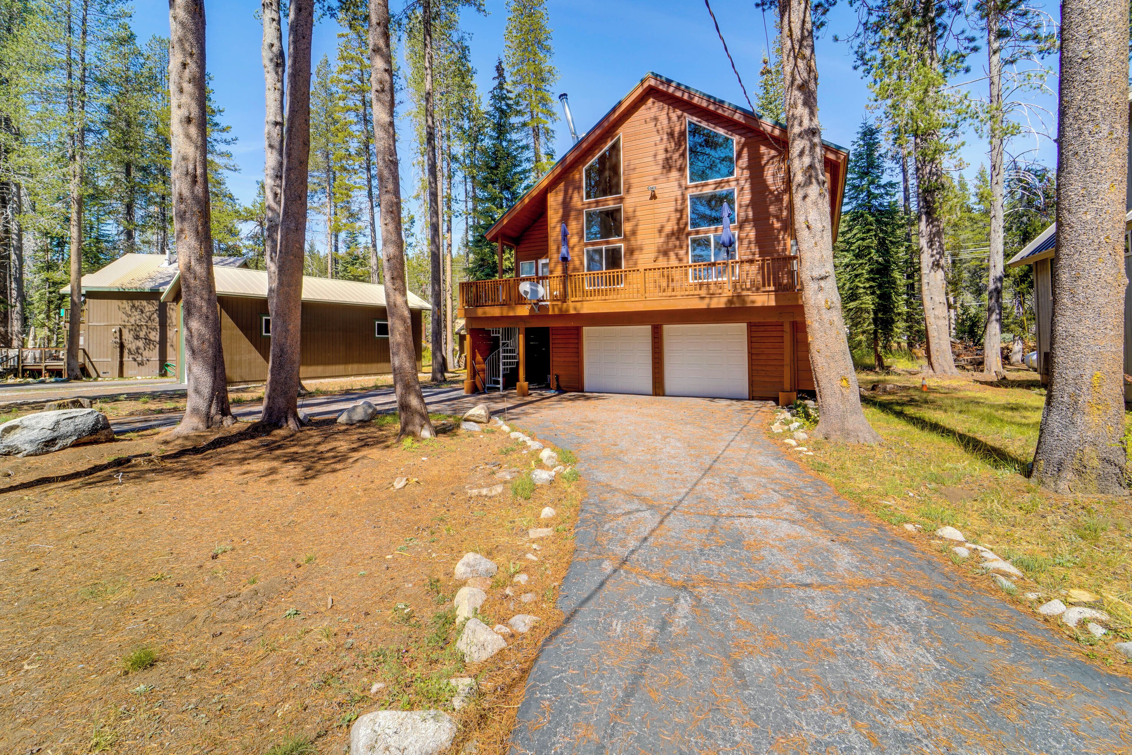 A two-story wooden cabin with a garage, surrounded by tall pine trees.