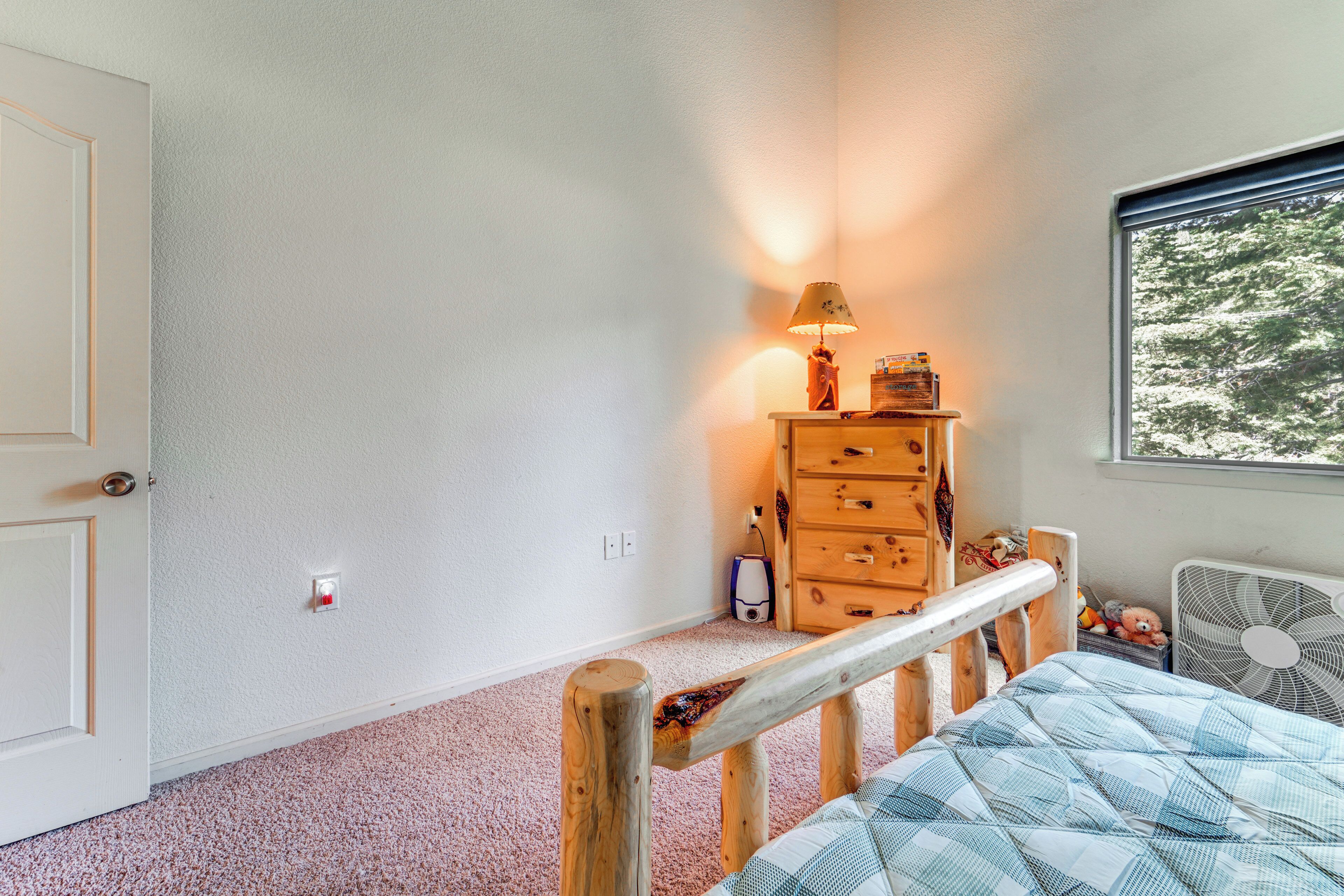 Ein Schlafzimmer mit einem hölzernen Kopfende, einem Bett mit einer blau-weißen Karodecke, einer Holzkommode, einer Lampe und einem Fenster mit Blick auf Bäume.