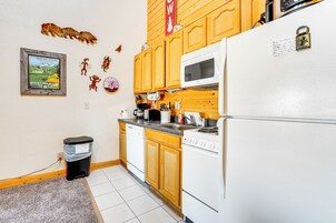 A kitchen with wooden cabinets, a white refrigerator, a microwave, and a stove. There is a trash can and a framed picture on the wall.