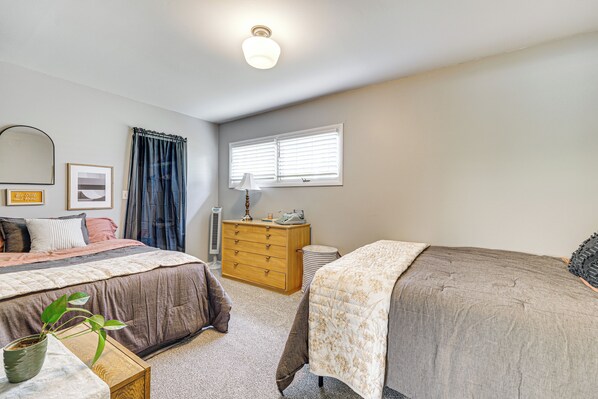 A bedroom with two beds, a dresser, a bedside table with a plant, a window with blinds, and a ceiling light. - Patio, Grill & Outdoor Dining: Conrad Home! (Conrad)