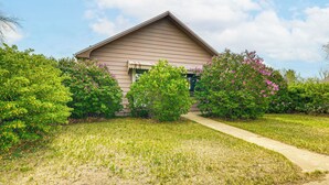 A single-story house with a sloped roof, surrounded by a well-maintained lawn and flowering bushes. - Patio, Grill & Outdoor Dining: Conrad Home! (Conrad)
